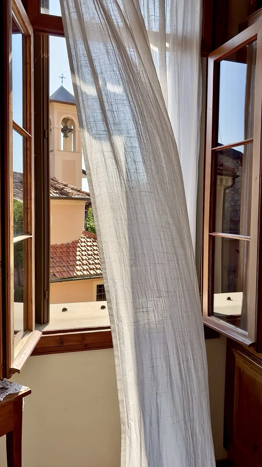 Open wooden window, white curtain and bell tower seen from Villa Panigai